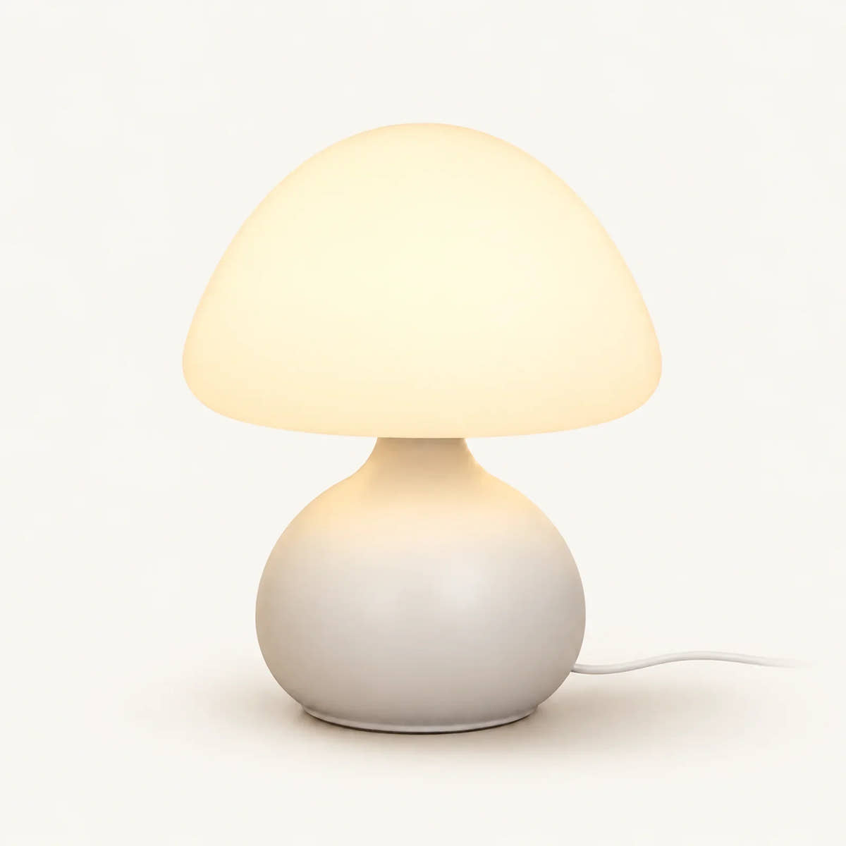 Cream mushroom table lamp with warm glowing dome shade, rounded base and white power cord on an off-white background.